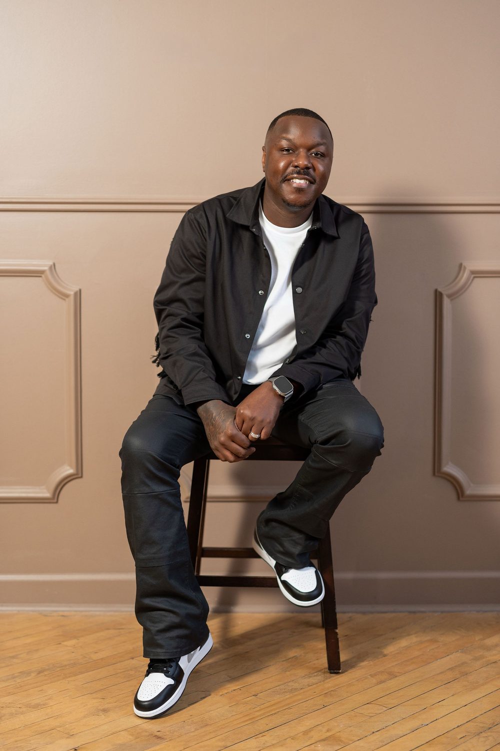 Jamel Lewis Sr., founder of The Brand Well, smiling and seated on a stool in a black jacket and white tee